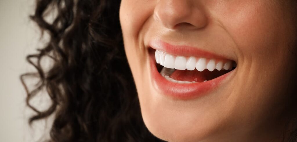 A close-up of a smiling woman with healthy, bright white teeth and naturally curly hair. The image emphasizes excellent oral hygiene, cosmetic dentistry, teeth whitening, and a confident, radiant smile.