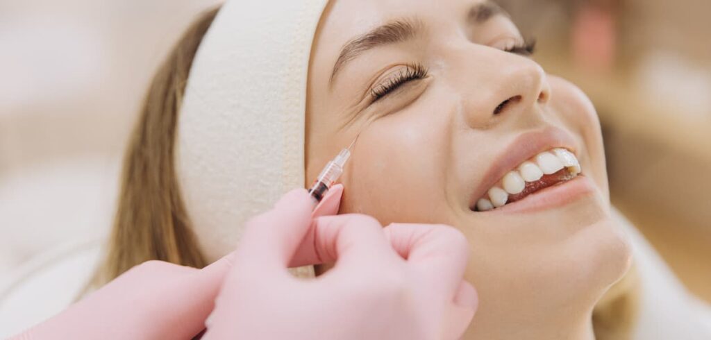 A patient smiles while receiving a cosmetic Botox injection near the outer corner of the eye from a qualified medical professional wearing gloves. The treatment is designed to reduce crow's feet and soften fine lines for a refreshed, youthful appearance.