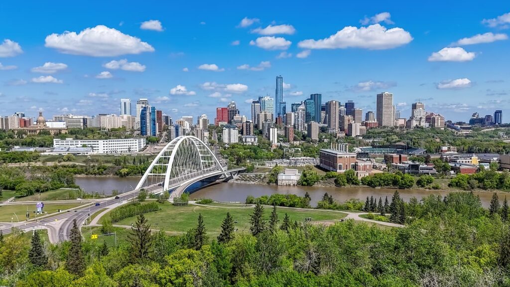 A panoramic view of downtown Edmonton featuring the Walterdale Bridge spanning the North Saskatchewan River. The city skyline rises above lush green parkland under a bright blue sky with scattered clouds, showcasing one of Alberta's most recognizable urban landscapes.