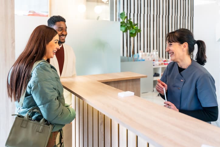A dental receptionist smiles while speaking with two patients at the front desk of a modern dental clinic. The bright reception area features contemporary wood accents and a welcoming atmosphere, highlighting a positive patient check-in experience.