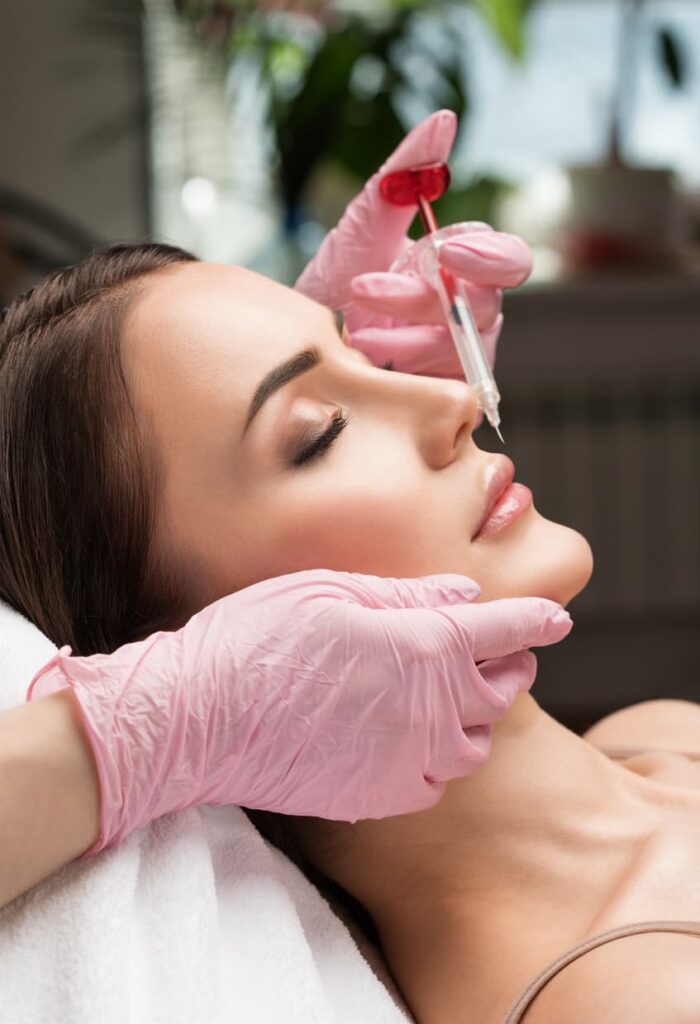 A medical aesthetics professional wearing pink gloves prepares to administer a lip filler injection while supporting the patient's chin. The minimally invasive cosmetic treatment is designed to enhance lip volume, improve symmetry, and create natural-looking results.