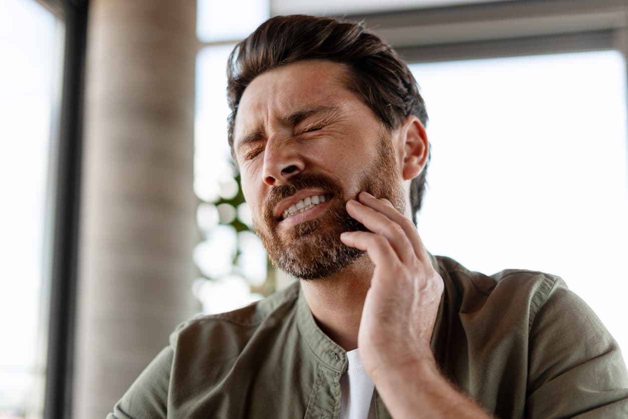 A man grimaces in pain while holding his jaw, appearing to suffer from a severe toothache or dental pain. The image represents common symptoms of a dental emergency, including tooth pain, infection, or sensitivity requiring prompt treatment.