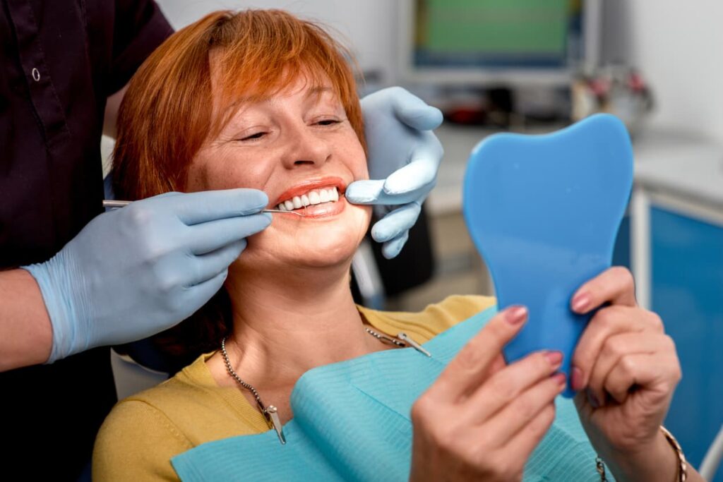 A smiling patient holds a handheld mirror while a dental professional checks the appearance of her teeth after cosmetic dental treatment. The image highlights smile enhancement, cosmetic dentistry, and patient satisfaction with treatment results.