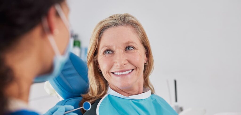 A smiling middle-aged patient wearing a dental bib sits in a dental chair while speaking with a dentist holding a dental mirror. The friendly interaction in a modern dental clinic highlights patient communication, comfort, and personalized dental care before treatment.