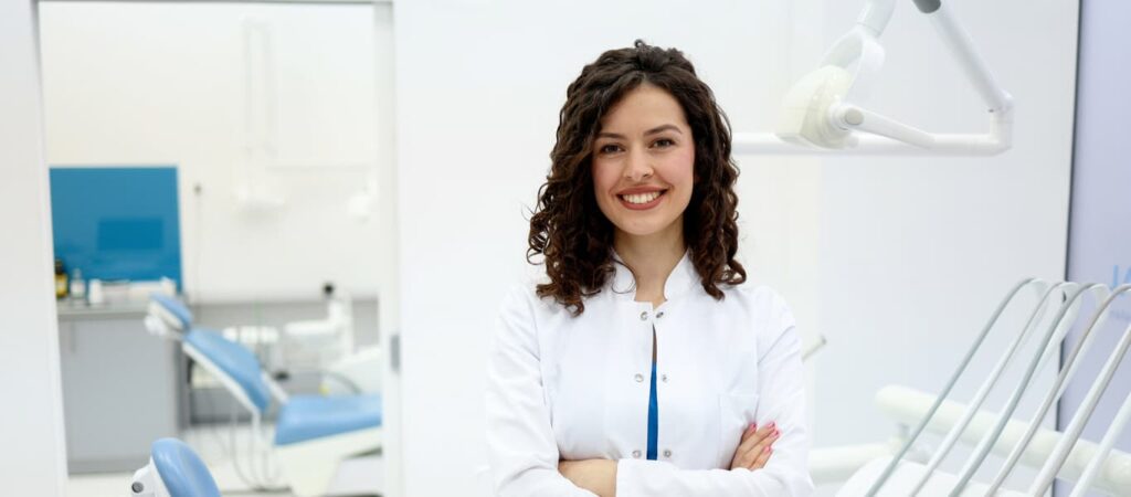 A smiling female dentist stands with her arms crossed in a bright, modern dental treatment room. Dental chairs and equipment are visible in the background, highlighting a professional, welcoming environment for oral healthcare.