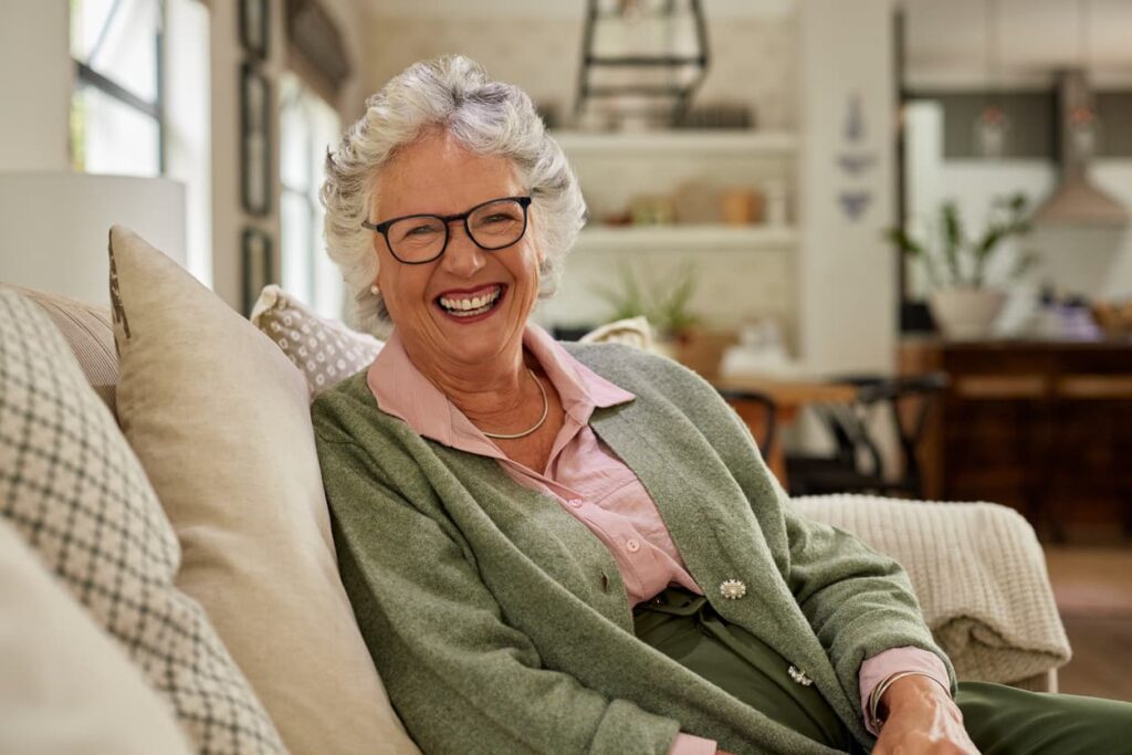 A smiling senior woman sits comfortably on a sofa at home, showing a healthy, confident smile. The image represents good oral health, preventive dental care, and maintaining a strong smile through every stage of life.