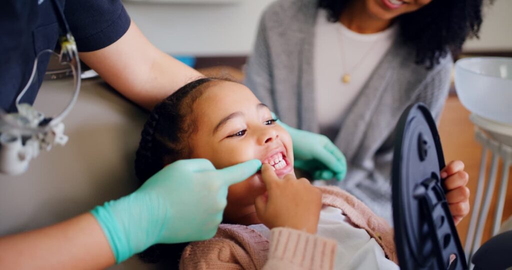 A dentist shows a young child their teeth using a handheld mirror during a pediatric dental appointment while a parent looks on. The image represents children's dentistry, preventive dental care, oral hygiene education, and family-focused dental visits.