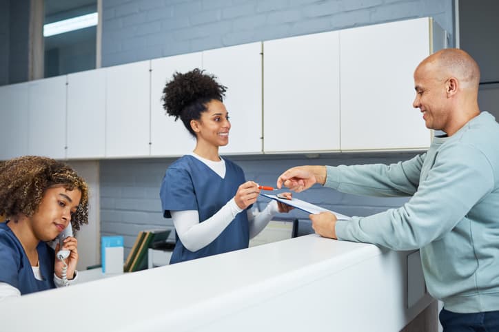 A dental receptionist greets a patient at the front desk while another team member answers the phone. The image represents a welcoming dental clinic, patient check-in, appointment scheduling, and friendly customer service.