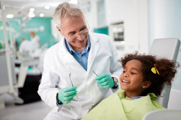 A dentist smiles and talks with a young girl before her dental examination, helping her feel relaxed and comfortable. The image represents children's dentistry, compassionate pediatric dental care, preventive checkups, and creating positive experiences for young patients.