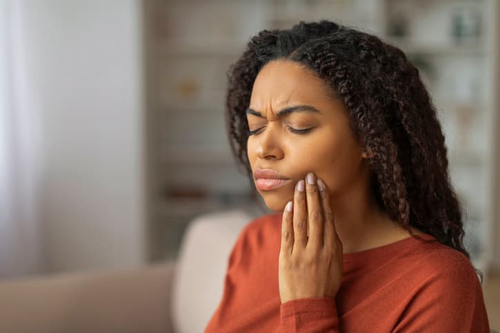 A woman holds her jaw with a pained expression, indicating a toothache or dental pain. The image highlights common symptoms of oral health issues such as tooth decay, infection, gum disease, or a dental emergency.