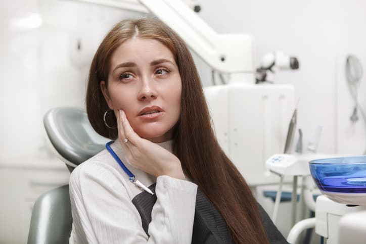 A woman sits in a dental chair holding her jaw in discomfort, showing signs of a painful toothache. The image represents dental pain caused by tooth decay, infection, an abscess, or another dental emergency that requires prompt assessment and treatment.