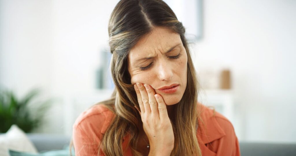 A woman holds her cheek with a painful expression, showing signs of a severe toothache or dental pain. The image represents common symptoms of tooth decay, infection, an abscessed tooth, or another dental emergency requiring prompt treatment.