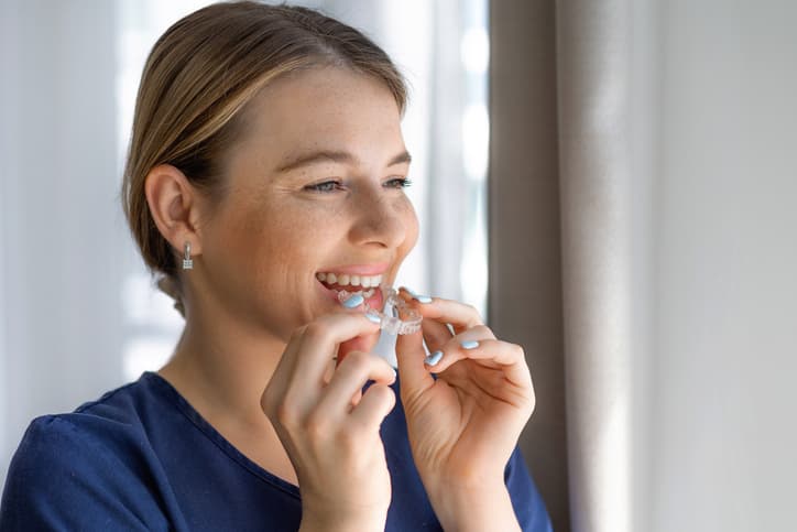 Smiling woman placing a clear dental aligner over her teeth, representing discreet orthodontic treatment for gradually straightening teeth and improving smile alignment.