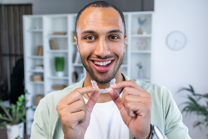 Smiling man holding a clear dental aligner in front of his teeth, representing discreet orthodontic treatment for improving tooth alignment and achieving a straighter smile.