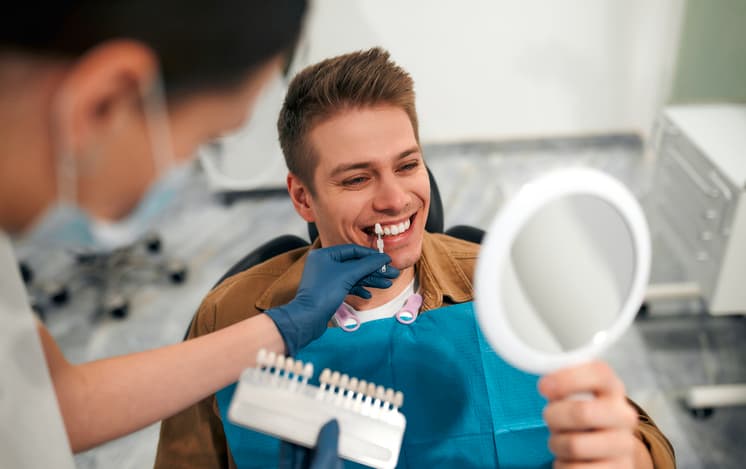 Smiling man looking in a mirror while a dental professional compares tooth shades during a cosmetic dentistry consultation.